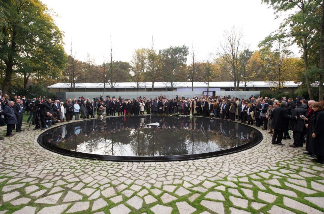 A large group of people stands at the edge of a round, black pool of water. The basin has a diameter of several meters. In the middle of the basin is flat black triangle, which is not covered by water. On it is placed a single white flower.